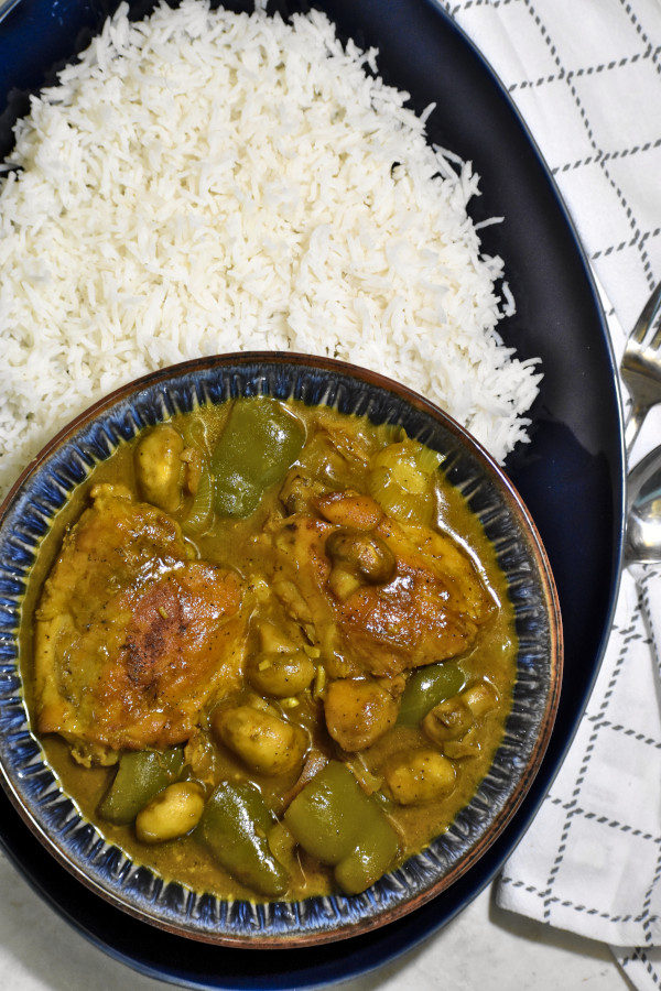 overhead shot of a blue bowl full of south african chicken curry on a large blue platter with rice. to the right is a white napkin and spoon and fork.