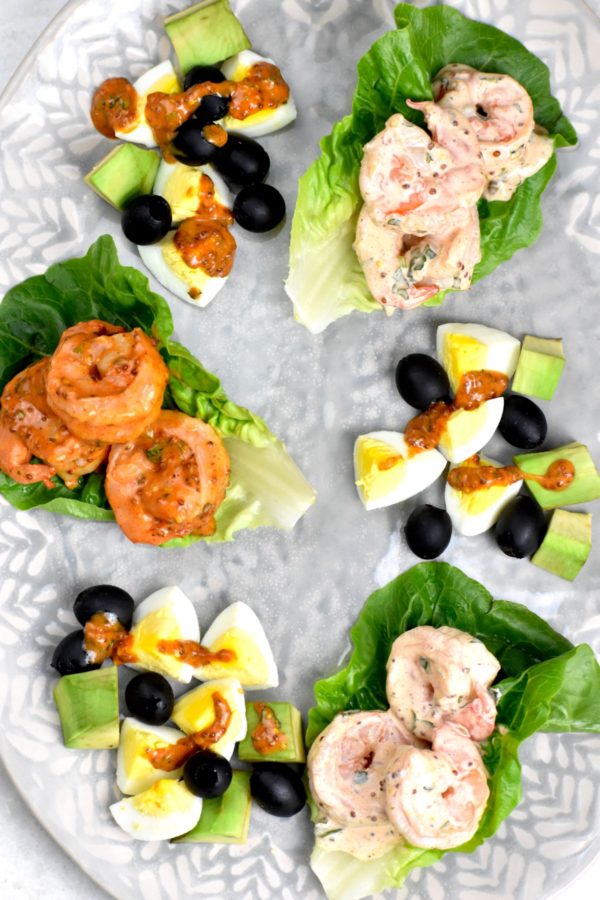 Three shrimp remoulade lettuce cups on a platter, two using the pink version of the sauce and one using the red, alongside garnishings of olives, hard boiled eggs and avocado.